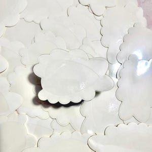 May include: A collection of white, cloud-shaped stickers with scalloped edges. The stickers are scattered across a pink surface, creating a textured, layered effect. The stickers are blank and ready for use.