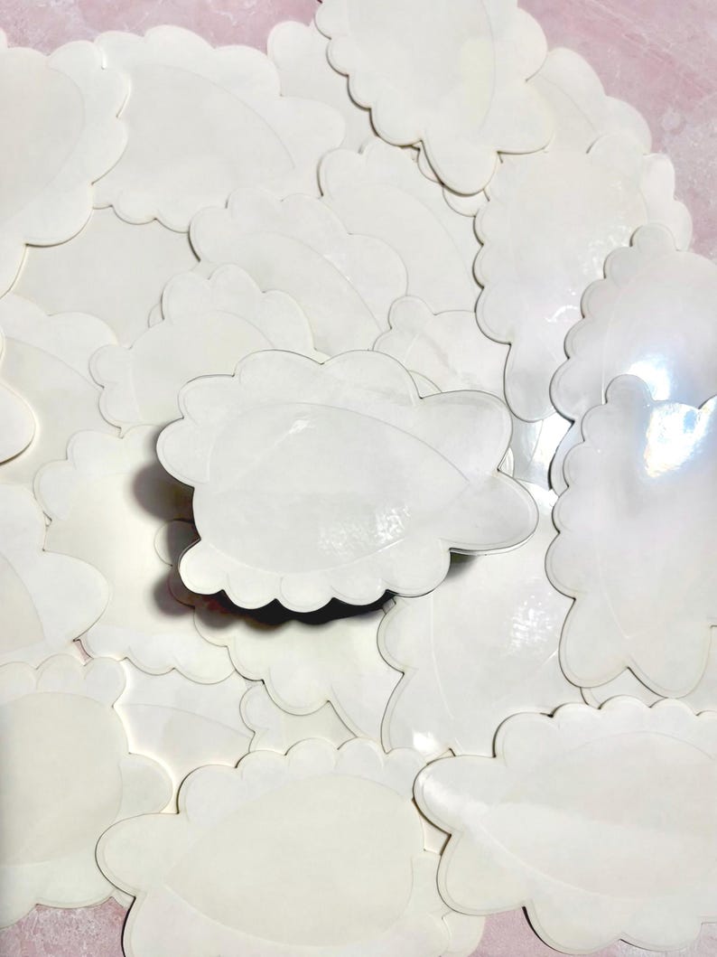 May include: A collection of white, cloud-shaped stickers with scalloped edges. The stickers are scattered across a pink surface, creating a textured, layered effect. The stickers are blank and ready for use.