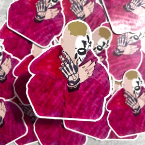 Könnte beinhalten: Eine Sammlung von gestanzten Aufklebern mit einer Person mit schädelähnlichem Gesicht und Skelett-Handtattoos. Die Person trägt einen roten Batik-Hoodie. Die Aufkleber haben einen weißen Rand.