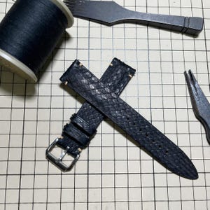 May include: A black watch strap with a textured snakeskin pattern, featuring a silver-toned metal buckle and light-colored stitching. The strap is positioned diagonally, accompanied by a spool of black thread and a metal tool nearby.