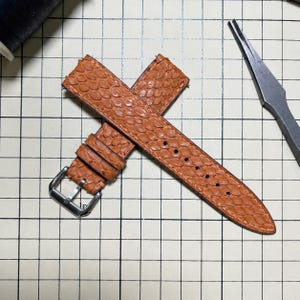 May include: A tan watch strap with a textured, scale-like pattern, featuring a silver-toned metal buckle and two matching leather keepers. The strap is positioned on a grid-lined surface next to a pair of metal pliers.