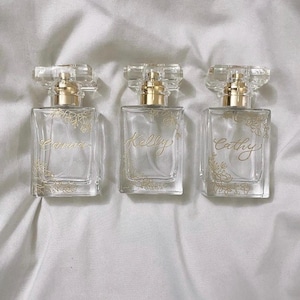 May include: Three clear glass perfume bottles with gold detailing and floral patterns. Each bottle has a gold cap and is labelled with a name: Carrie, Kelly, and Cathy. The bottles are arranged on a white, textured surface.