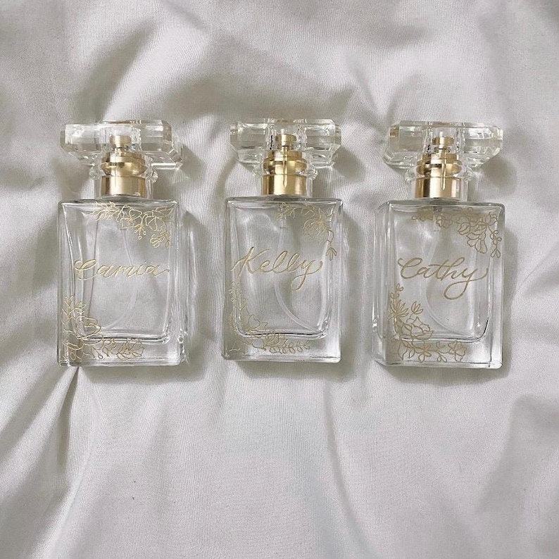 May include: Three clear glass perfume bottles with gold detailing and floral patterns. Each bottle has a gold cap and is labelled with a name: Carrie, Kelly, and Cathy. The bottles are arranged on a white, textured surface.