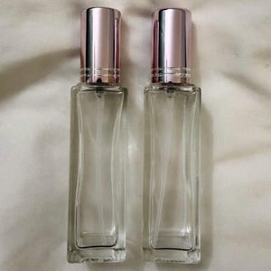 May include: Two clear glass perfume bottles with rose gold coloured caps. The bottles are rectangular with a slightly curved shape. The caps are cylindrical and have a shiny finish. The bottles are empty and appear to be new.