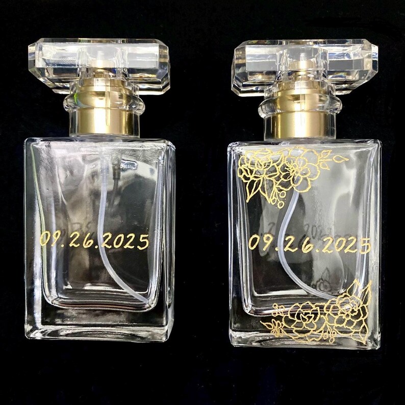 May include: Two clear glass perfume bottles with gold accents and crystal stoppers. One bottle has gold floral detailing and the date "09.26.2025" in gold. The other bottle has the same date. Both bottles are on a black background.