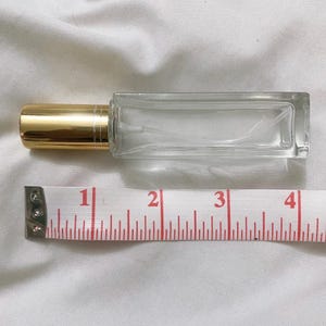 May include: A clear glass perfume bottle with a gold-coloured cap. The bottle is rectangular and measures approximately 10 cm in length. A measuring tape is placed next to the bottle.