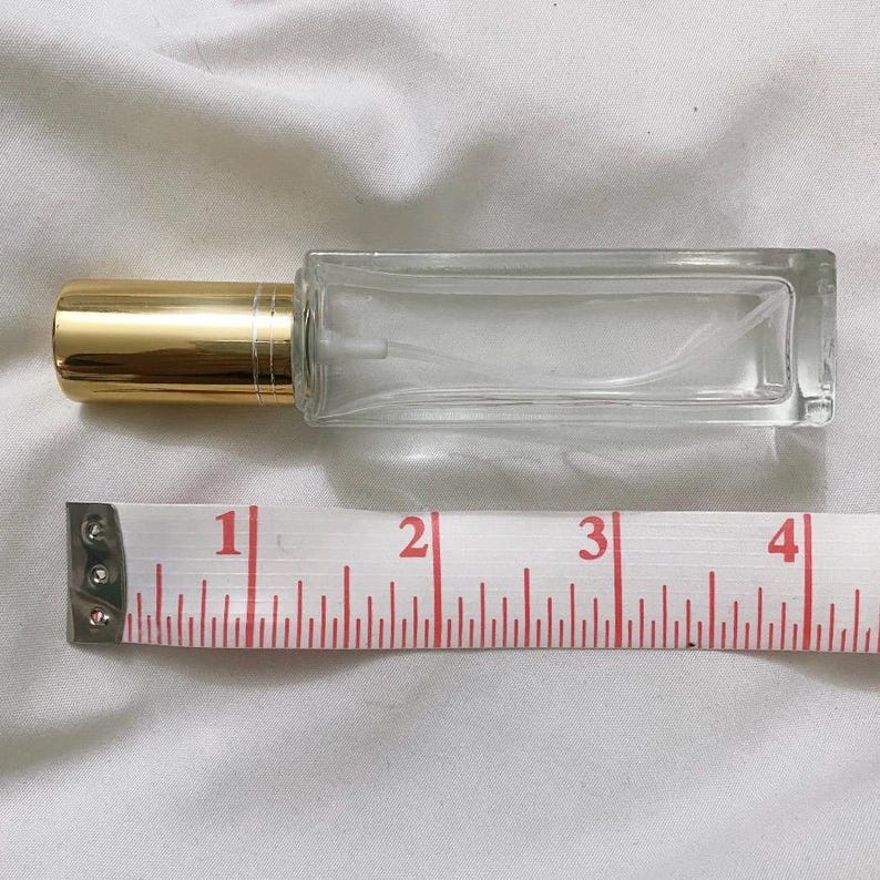 May include: A clear glass perfume bottle with a gold-coloured cap. The bottle is rectangular and measures approximately 10 cm in length. A measuring tape is placed next to the bottle.
