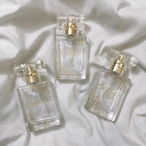 May include: Three clear glass perfume bottles with gold detailing and floral patterns. Each bottle is labelled with a different name in gold script: Kelly, Cathy, and Camia. The bottles are on a white, textured surface.