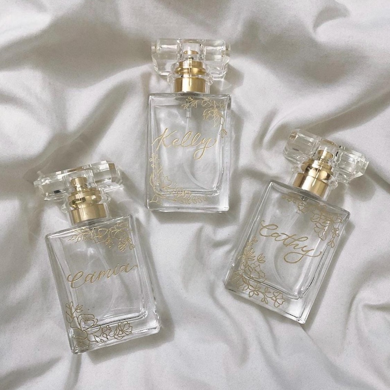 May include: Three clear glass perfume bottles with gold detailing and floral patterns. Each bottle is labelled with a different name in gold script: Kelly, Cathy, and Camia. The bottles are on a white, textured surface.