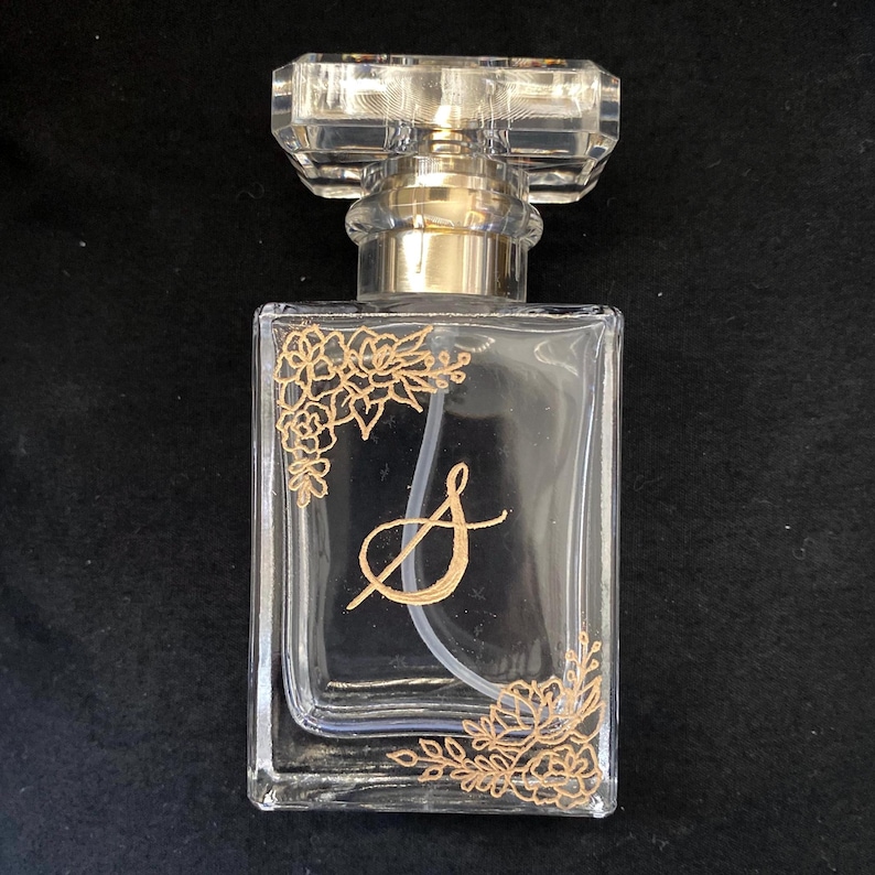 May include: Clear glass perfume bottle with a rectangular shape and a faceted, clear stopper. The bottle features gold floral designs and a gold letter "S". The atomizer is gold.