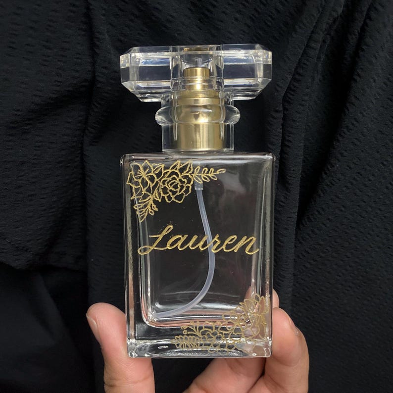 May include: A clear glass perfume bottle with a gold-coloured sprayer and a clear, rectangular cap. The bottle is adorned with gold floral designs and the name "Lauren" in elegant script. The bottle is held against a dark background.