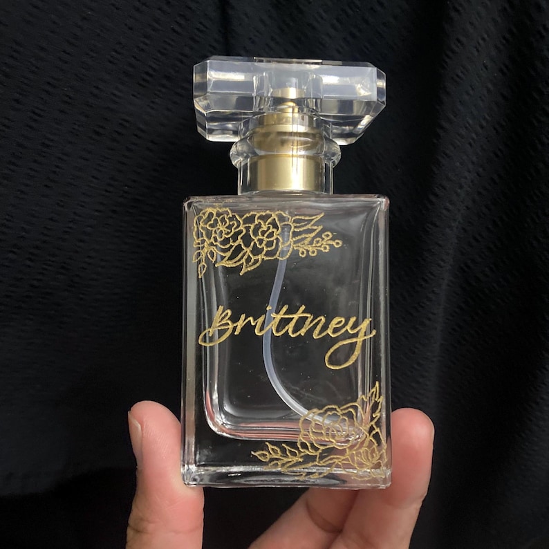 May include: A clear glass perfume bottle with a rectangular shape and a clear, faceted cap. The bottle is decorated with gold floral designs and the name "Brittney" in gold script. The bottle is held against a dark background.