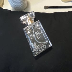 May include: A clear perfume bottle with a rectangular shape and a crystal-like cap. The bottle is decorated with floral designs and the name "Cathy" in elegant script. The bottle has a gold-coloured neck.
