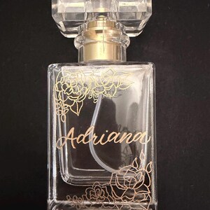 May include: A clear glass perfume bottle with a gold-coloured atomizer and a faceted, rectangular cap. The bottle is decorated with gold floral designs and the name "Adriana" in elegant script. The background is black.