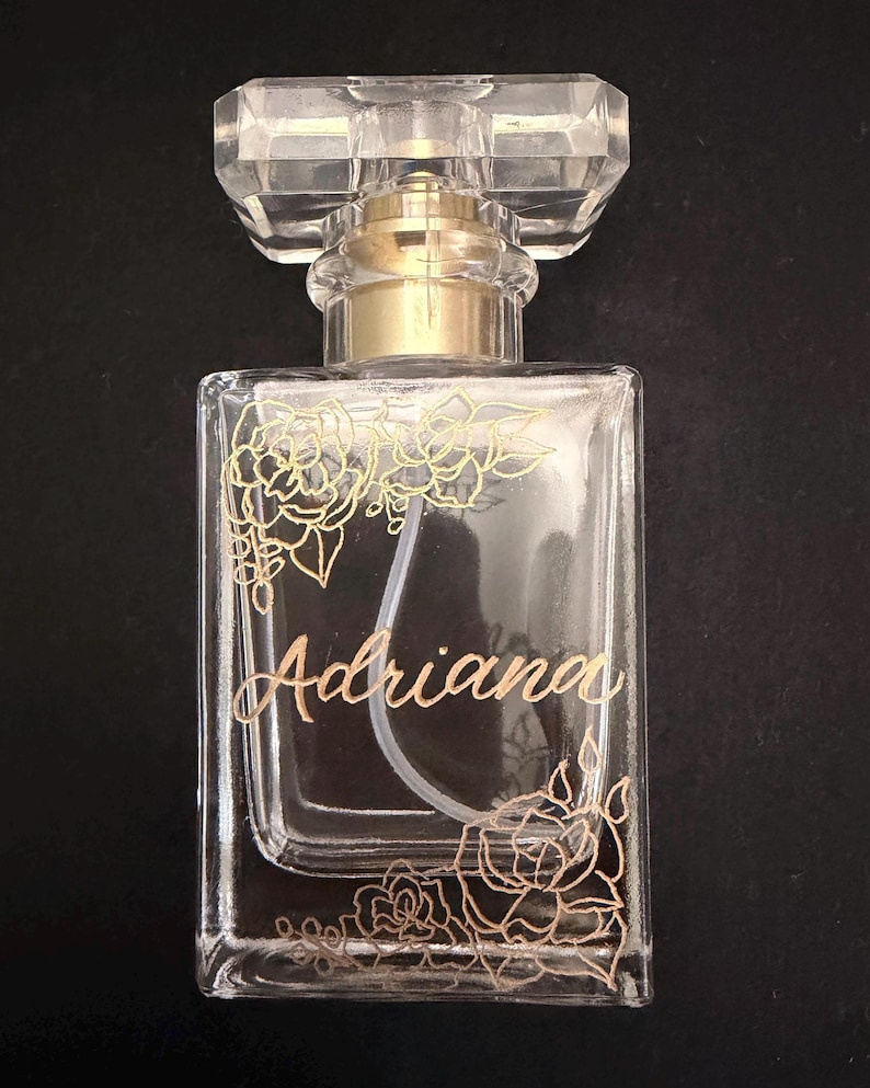 May include: A clear glass perfume bottle with a gold-coloured atomizer and a faceted, rectangular cap. The bottle is decorated with gold floral designs and the name "Adriana" in elegant script. The background is black.