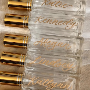 May include: Clear glass perfume bottles with gold-coloured caps. Each bottle is labelled with a name in gold script: Katelyn, Katie, Kennedy, Megan, Lindsey, Katlynn, and Brooke. The bottles are arranged in a row.