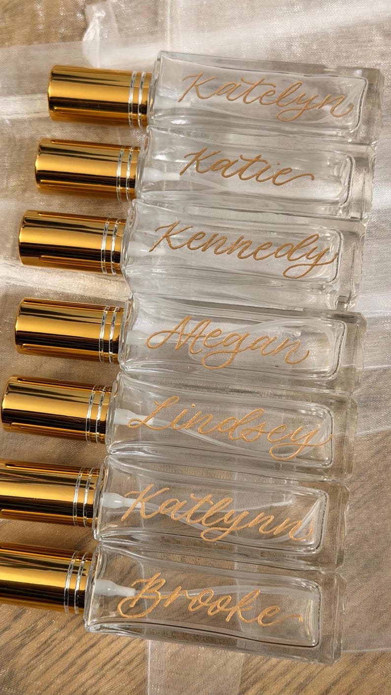 May include: Clear glass perfume bottles with gold-coloured caps. Each bottle is labelled with a name in gold script: Katelyn, Katie, Kennedy, Megan, Lindsey, Katlynn, and Brooke. The bottles are arranged in a row.
