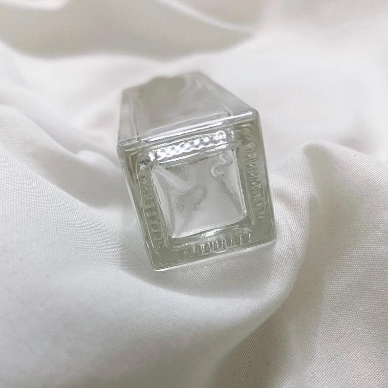 May include: A clear, rectangular glass bottle with a square base. The bottle has a slightly textured surface and is set against a white, wrinkled fabric background. The bottle's design is simple and minimalist.
