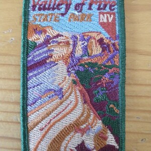 May include: A rectangular embroidered patch featuring a landscape of Valley of Fire State Park NV. The design includes red, orange, purple, and white rock formations under a light blue sky, all bordered by a thick dark green stitched edge.