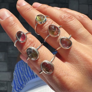 May include: A set of six silver rings featuring different coloured gemstones. The gemstones are oval shaped and come in a range of colours, including green, red, and brown.