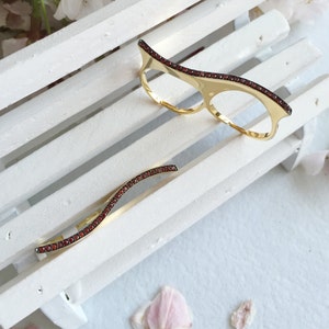 May include: Two gold rings with a wavy design and red gemstones. The rings are on a white wooden surface.