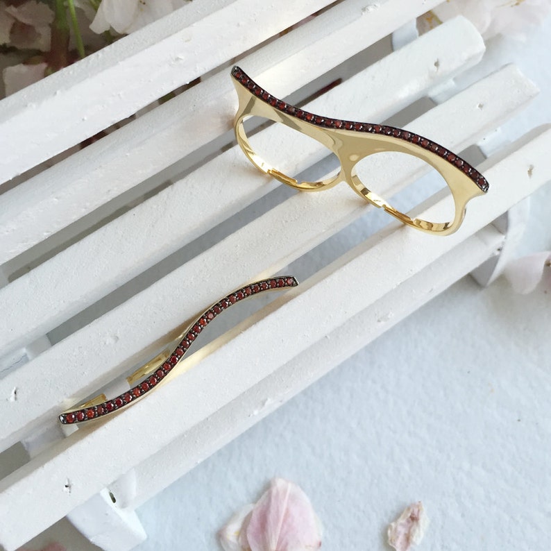 May include: Two gold rings with a wavy design and red gemstones. The rings are on a white wooden surface.