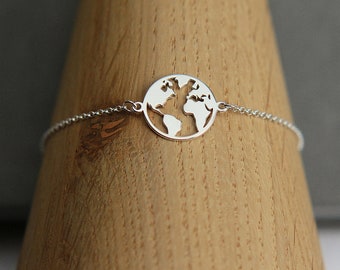 May include: A silver-colored bracelet featuring a circular pendant with a cut-out design of the Earth's continents. The bracelet's chain is delicate, and the pendant is approximately 1 inch in diameter. The bracelet is displayed on a wooden surface.