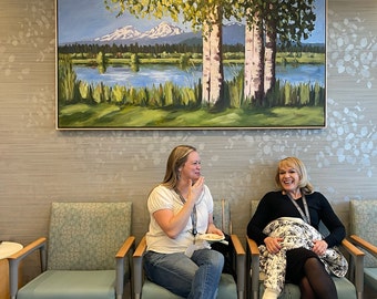 May include: Two people are sitting in chairs in front of a painting of a lake and mountains. The painting is in a light wood frame. The people are both smiling and looking at each other.