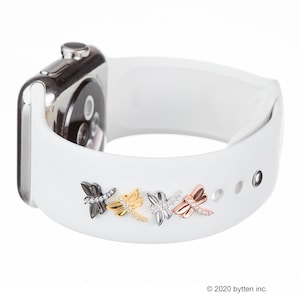 May include: A white Apple Watch band with four dragonfly charms in black, gold, silver, and rose gold. Each charm has clear rhinestones.