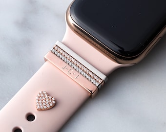 May include: A pink Apple Watch band with a rose gold finish. The band has a heart-shaped charm with clear rhinestones and a double row of rhinestones on the band. The band also has a personalized inscription that reads "J.C&J".