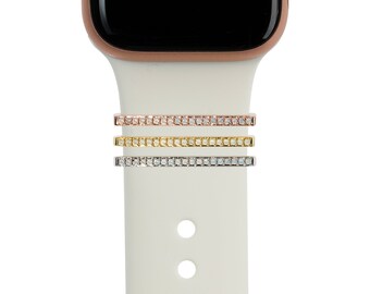 May include: Three watch band accessories in rose gold, gold, and silver, each adorned with a row of small, sparkling stones. The accessories are shown on a white watch band, with a rose gold watch face visible at the top.