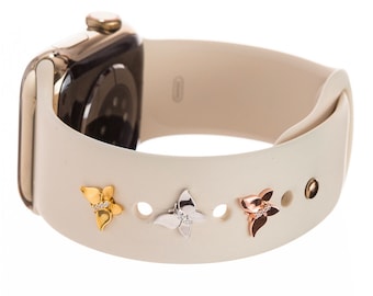 Pode incluir: Uma pulseira de relógio Apple de silicone branca com três pingentes decorativos. Os pingentes têm a forma de uma flor com uma única fila de pequenos diamantes. Os pingentes são de ouro, prata e ouro rosa.