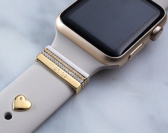 May include: A gold Apple Watch with a white band and a gold band with a row of small diamonds and the inscription "11.26.02".