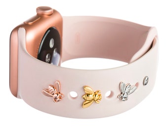 May include: A pink silicone Apple Watch band with three bee charms. The charms are gold, silver, and rose gold.