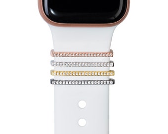 May include: Four decorative watch band charms in rose gold, silver, gold, and black, each encrusted with small, clear crystals. The charms are shown on a white watch band attached to a rose gold watch face. The watch band has three circular holes.