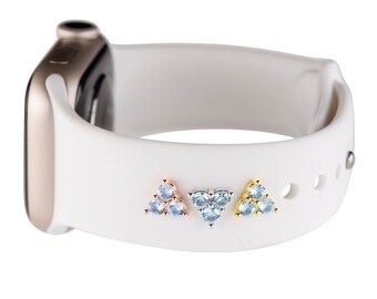 May include: A white Apple Watch band with three small triangle-shaped charms with blue gemstones. The charms are in different metal finishes: rose gold, silver, and gold.