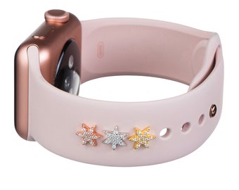 May include: A rose gold Apple Watch with a light pink silicone band. Three star-shaped charms with clear and gold rhinestones are attached to the band.