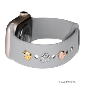 May include: A gray silicone Apple Watch band with three ladybug charms. The charms are gold, silver, and rose gold with clear rhinestones.
