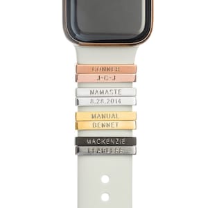 Può includere: Un cinturino bianco per Apple Watch con più ciondoli in metallo personalizzati. I ciondoli sono incisi con le parole "CONNER", "J*C*J", "NAMASTE", "8.28.2014", "MANUAL", "BENNET", "MACKENZIE" e "FEARLESS".