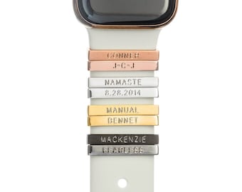 May include: A white Apple Watch band with multiple personalized metal charms. The charms are engraved with the words "CONNER", "J*C*J", "NAMASTE", "8.28.2014", "MANUAL", "BENNET", "MACKENZIE", and "FEARLESS".