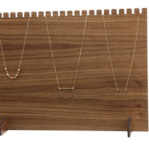 May include: A wooden necklace display stand with three gold necklaces hanging on it. The stand has a brown wood finish and a rectangular shape. The stand has two small feet on the bottom.