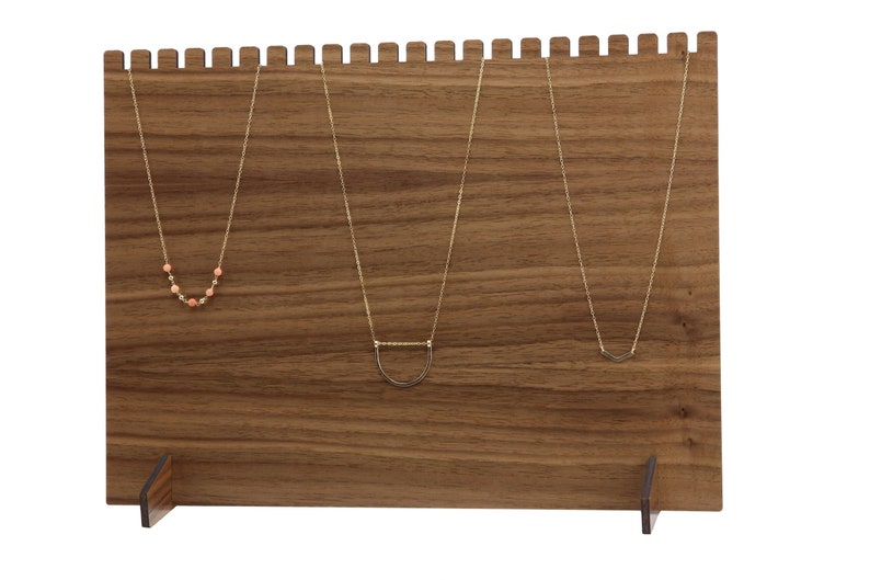 May include: A wooden necklace display stand with three gold necklaces hanging on it. The stand has a brown wood finish and a rectangular shape. The stand has two small feet on the bottom.