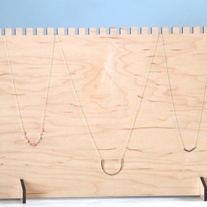 May include: A wooden necklace display stand with three necklaces hanging from it. The stand is made of light wood and has a simple design. The necklaces are all different styles, with one featuring small pink beads.