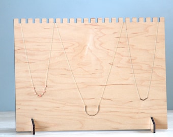 May include: A wooden necklace display stand with three necklaces hanging from it. The stand is made of light wood and has a simple design. The necklaces are all different styles, with one featuring small pink beads.