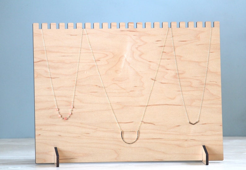 May include: A wooden necklace display stand with three necklaces hanging from it. The stand is made of light wood and has a simple design. The necklaces are all different styles, with one featuring small pink beads.