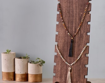 May include: A dark brown wooden necklace display stand with two necklaces hanging on it. The necklaces are made of colorful beads and have tassels at the bottom. The stand has a scalloped top and a base with two feet.