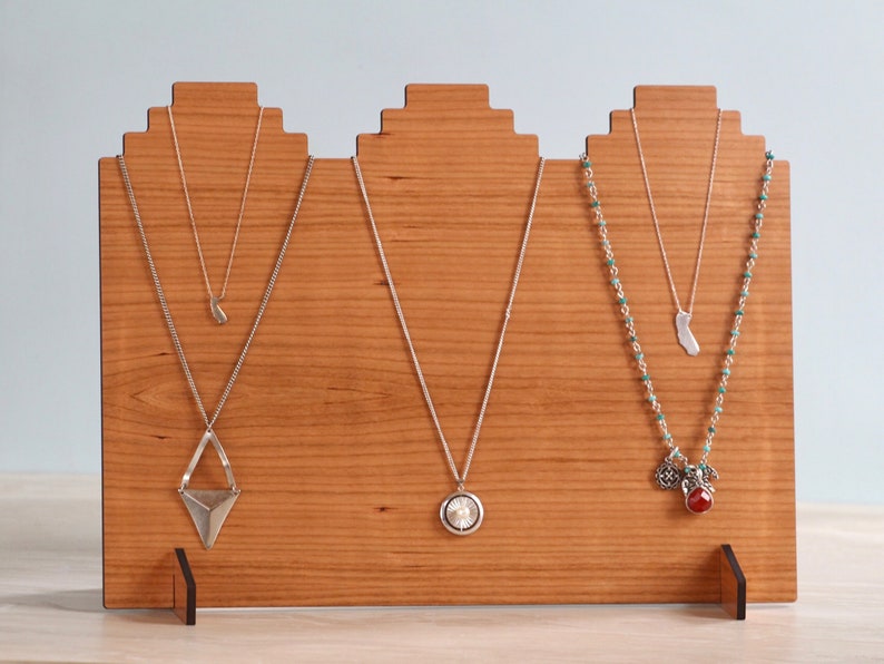 May include: A wooden necklace display stand with three slots. Each slot has a different necklace with a silver pendant. The necklaces are on a light brown wooden background.