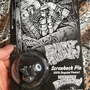 Peut inclure: Une épingle à revers en argent en forme de moteur de moto, emballée dans une bulle en plastique transparente sur une carte noire avec le texte "Gnarly Pounder! Screwback Pin 100% Recycled Pewter! Seriously Disgusting! Series #1 2024"