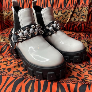 Puede incluir: Un par de botines blancos brillantes con suelas negras y paneles laterales elásticos. Las botas tienen una correa negra adornada con púas plateadas y cuentas blancas. Las suelas gruesas tienen un diseño tachonado.