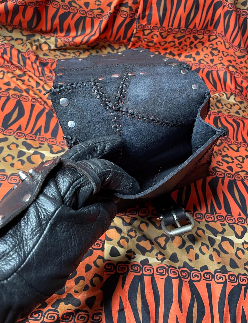 Pu&ograve; includere: Una borsa in pelle nera con chiusura a lembo e borchie argentate. La borsa &egrave; aperta e l'interno &egrave; foderato in tessuto nero. La borsa &egrave; appoggiata su un tessuto a fantasia.